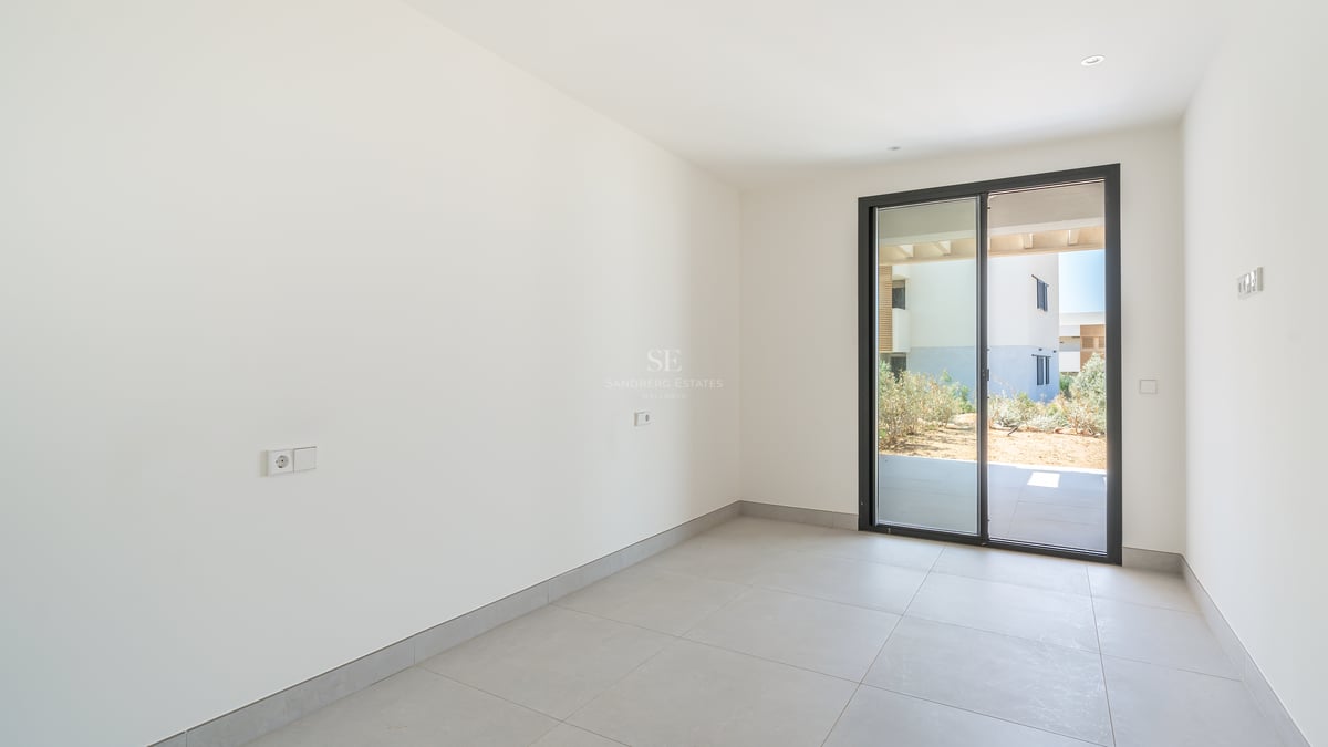 Dormitorio vacío con paredes blancas, suelo de gres gris y puerta corredera de cristal hacia un patio privado.