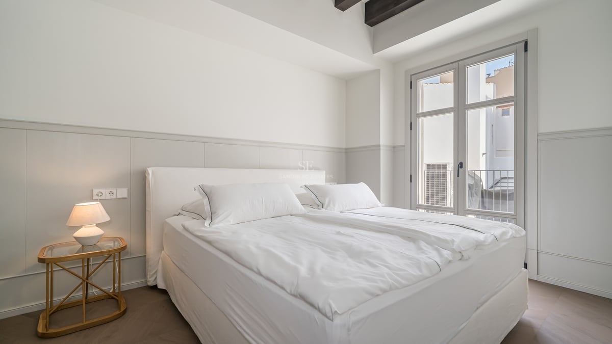 Dormitorio luminoso con cama blanca, vigas de madera oscura en el techo y ventanal hacia un balcón tipo Julieta.