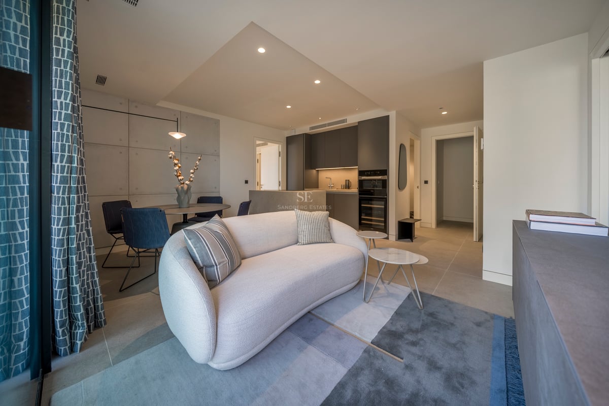 Salon moderne avec un canapé blanc incurvé, un tapis bleu et une table à manger, à côté d'une cuisine gris foncé.