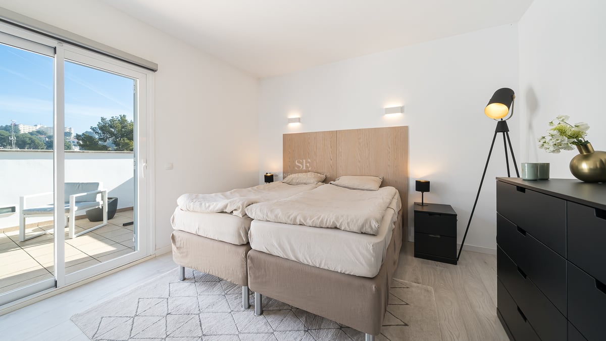 Chambre lumineuse avec un lit double, une tête de lit en bois clair et une baie vitrée ouvrant sur une terrasse ensoleillée.