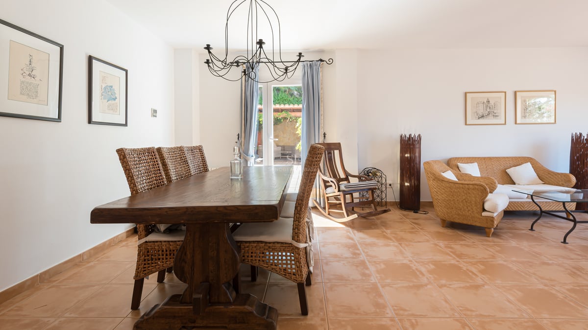 Salle à manger avec table en bois massif, chaises en osier et sol en terre cuite, s'ouvrant sur un coin salon.