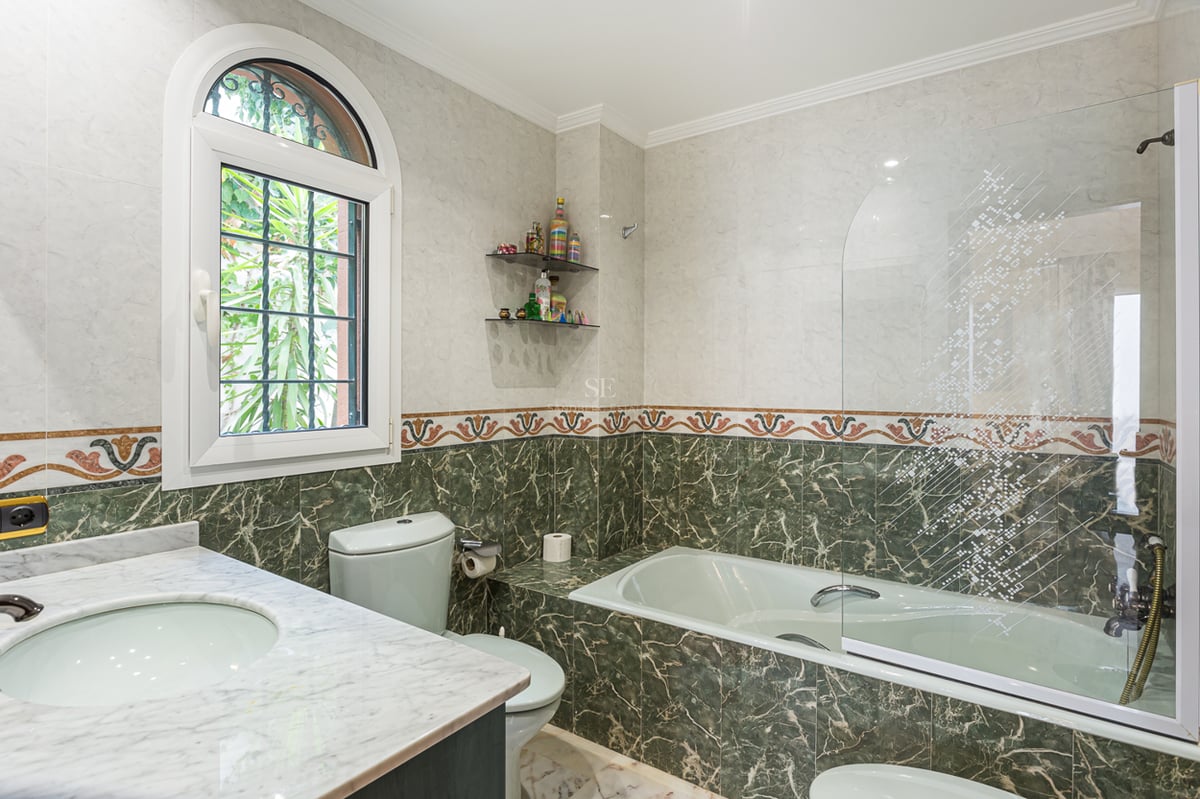 Salle de bain avec carrelage en marbre vert et blanc, baignoire avec paroi en verre et fenêtre cintrée.