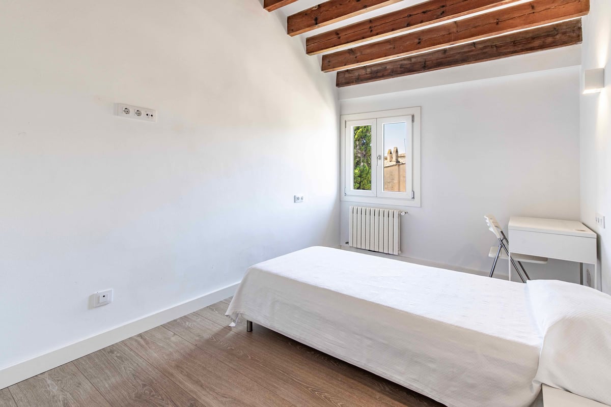 Chambre blanche avec lit simple, poutres en bois sombre au plafond incliné et parquet clair.