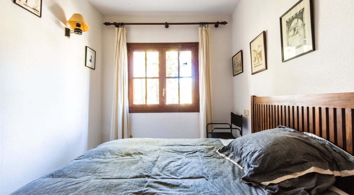 Une chambre avec un lit en bois, une couette grise et une fenêtre laissant passer une vive lumière du soleil.