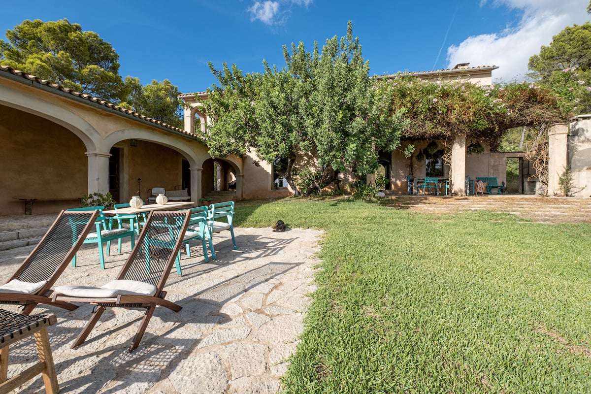 Un jardin ensoleillé avec patio en pierre, pelouse, chaises bleu sarcelle et une villa rustique aux arcades.