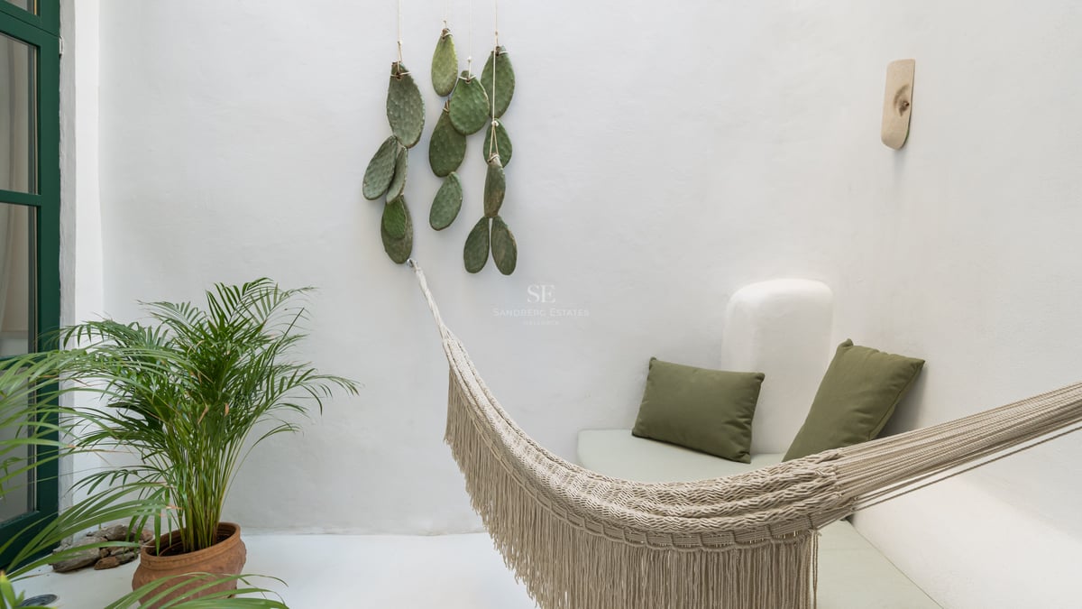 Patio aux murs blancs avec hamac à franges, coussins vert olive et plantes tropicales.