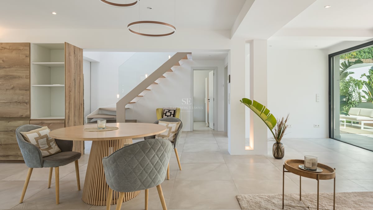 Salle à manger contemporaine avec table ronde en bois, chaises grises et escalier suspendu près des baies vitrées.