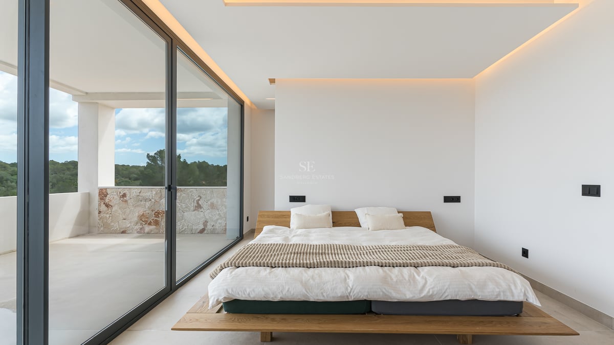 Chambre principale minimaliste avec lit en bois, baies vitrées et accès à un balcon en pierre naturelle.