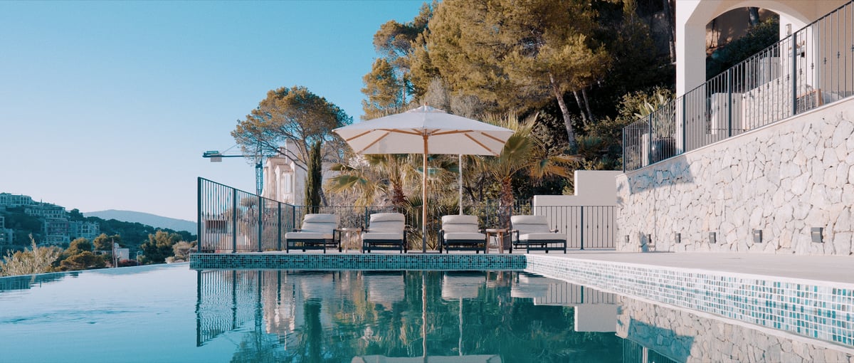 Piscine à débordement avec quatre chaises longues sous un parasol blanc, entourée de murs en pierre et de pins.