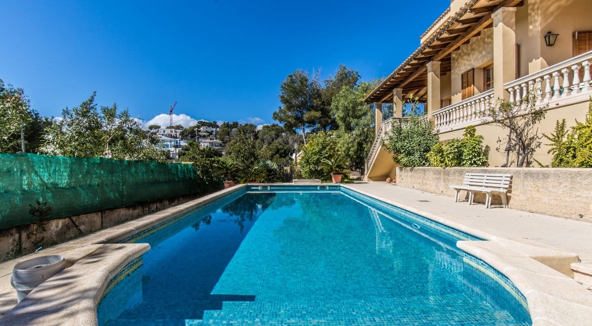 Grande piscine rectangulaire aux eaux turquoises à côté d'une villa méditerranéenne avec terrasse ensoleillée et ciel bleu.