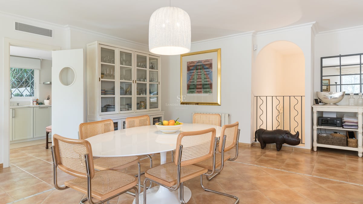 Salle à manger lumineuse avec table ovale blanche, chaises en rotin, sol en terre cuite et grand vaisselier.