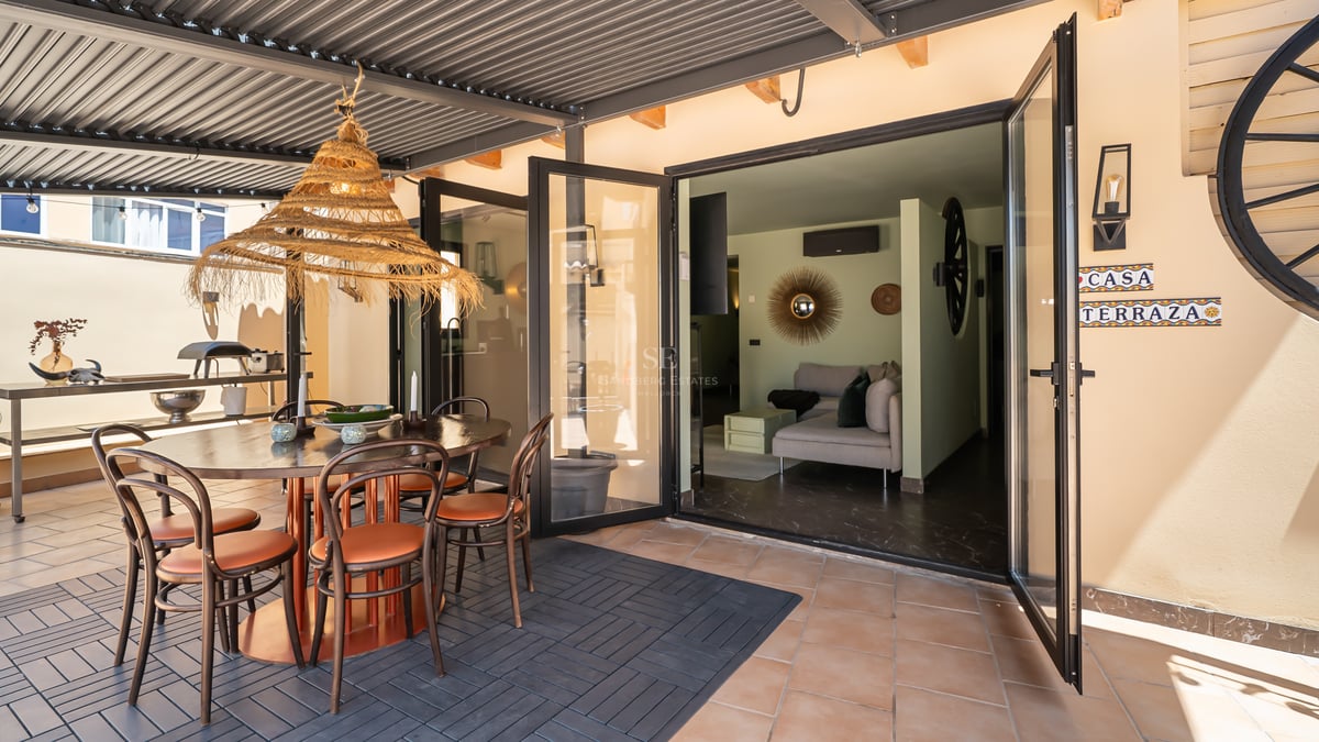 Une terrasse couverte avec une table à manger ronde, une suspension en rotin et des baies vitrées ouvrant sur le salon.