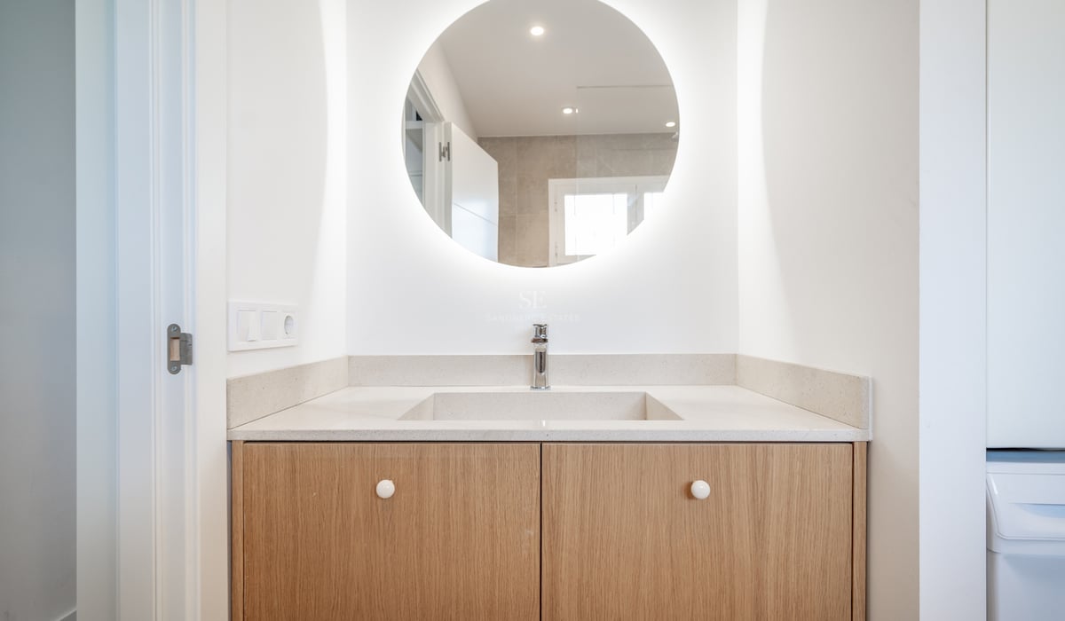 Vanité de salle de bain moderne avec miroir LED circulaire, ébénisterie en bois clair et lavabo blanc.