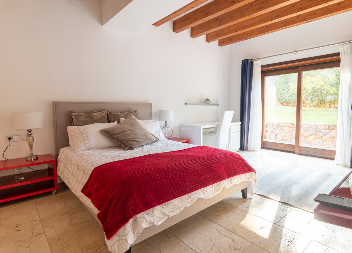 Chambre lumineuse avec accents rouges, sol en pierre, poutres en bois et baies vitrées donnant sur le jardin.