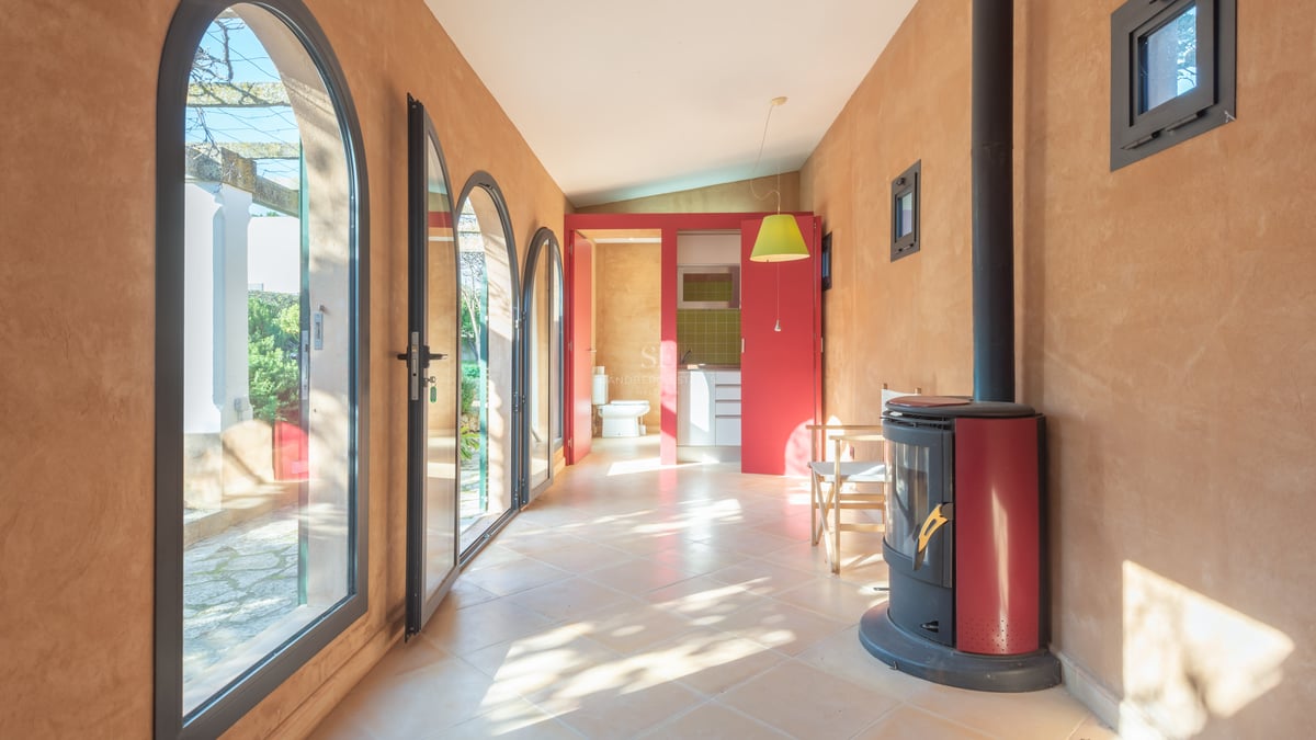 Pièce ensoleillée avec murs en terre cuite, portes vitrées cintrées, poêle à bois rouge et vue sur le jardin.