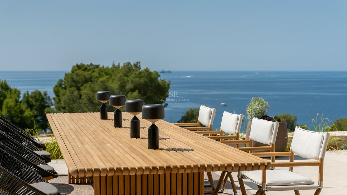 Table à manger moderne en bois avec lampes design sur une terrasse ensoleillée surplombant la Méditerranée.