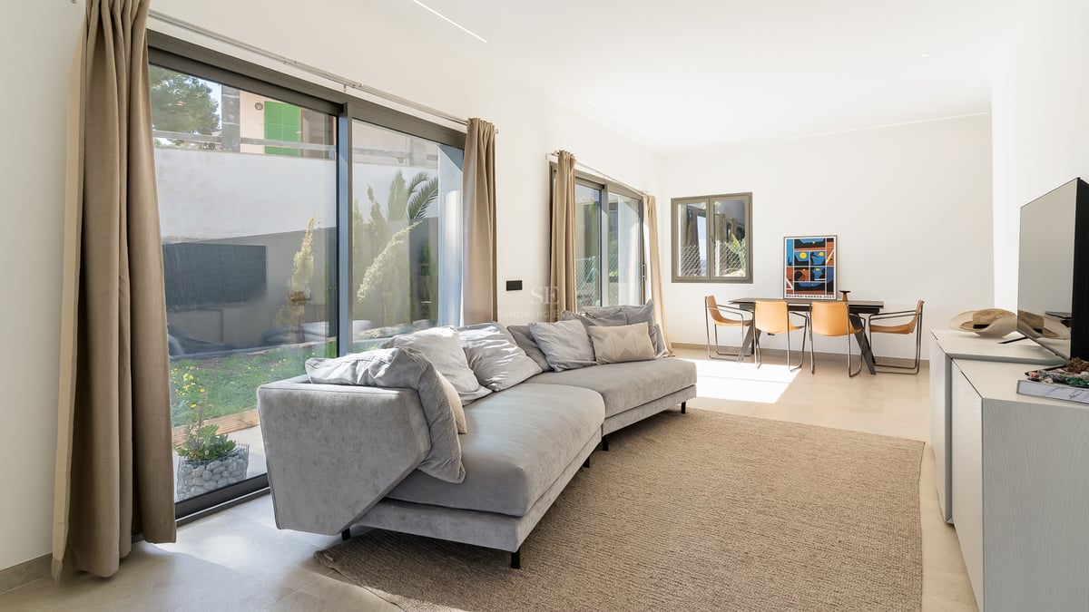 Salon lumineux avec grand canapé gris, baies vitrées donnant sur le jardin et coin repas avec chaises en cuir.