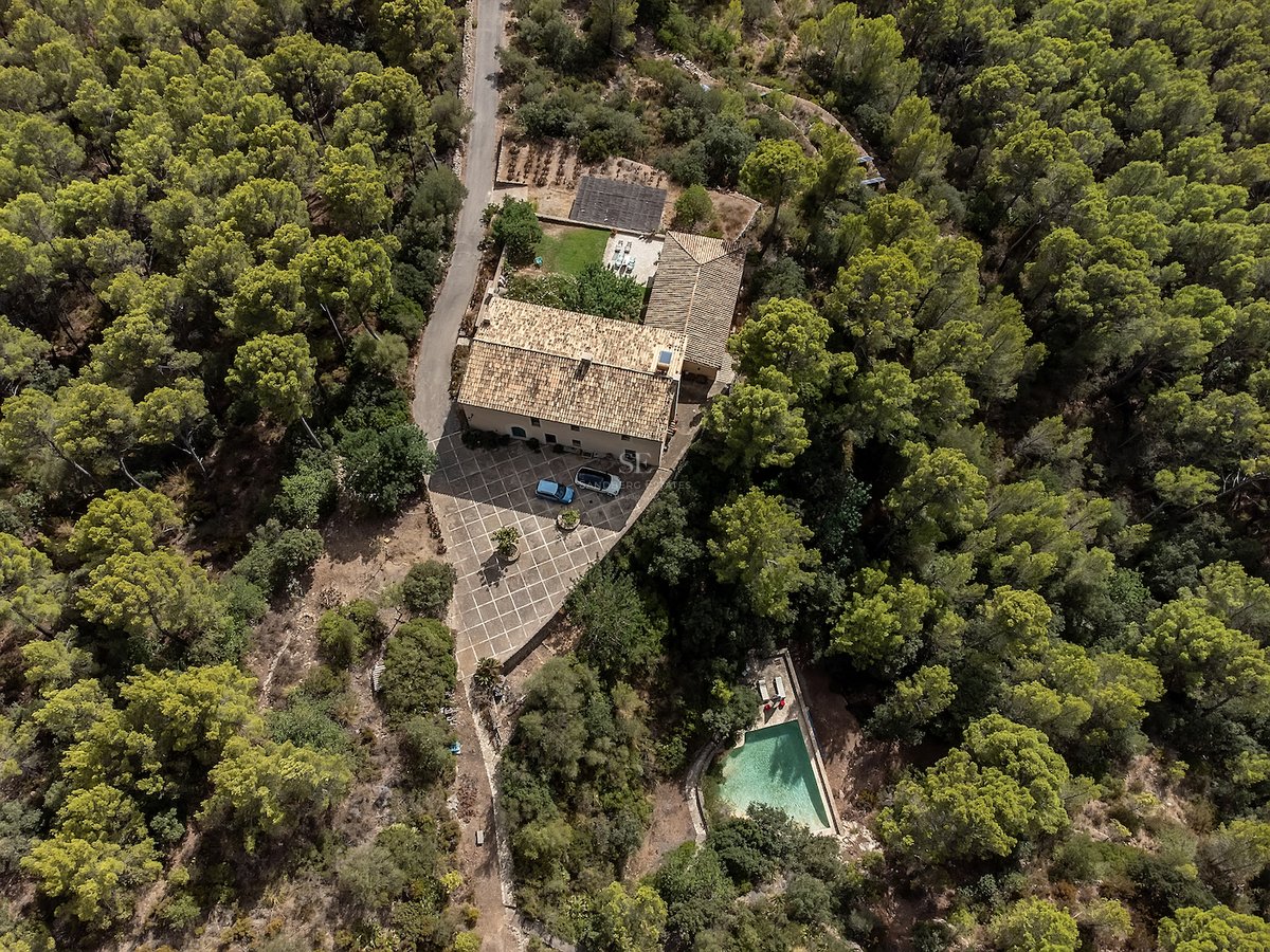 Vue aérienne d'une maison en pierre avec toit en terre cuite, cour et piscine, entourée d'une forêt de pins dense.