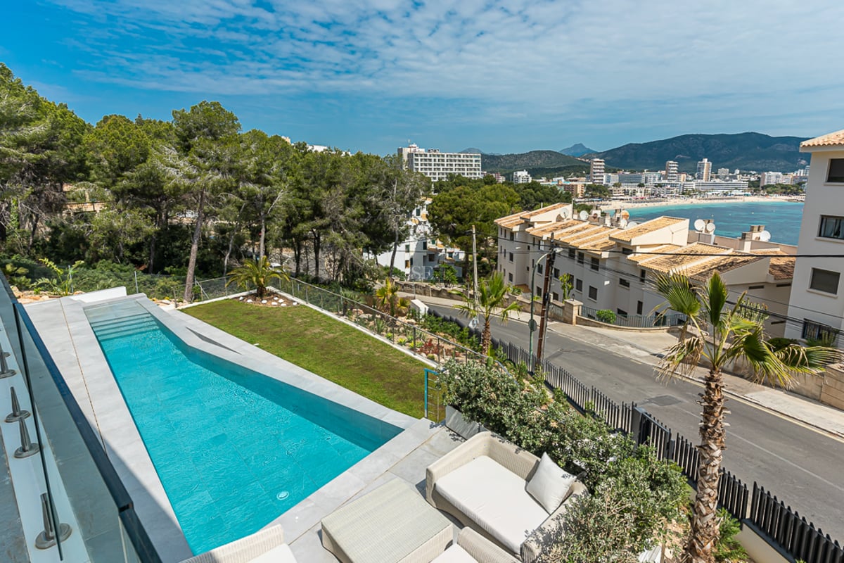 Une longue piscine turquoise sur une terrasse avec garde-corps en verre, surplombant une ville côtière et des montagnes.