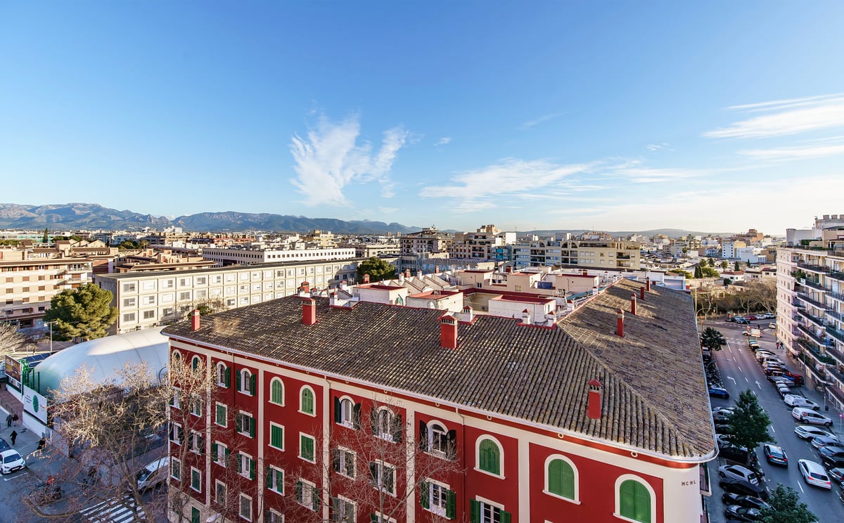Vue en plongée d'une ville avec un bâtiment rouge, des rues et des montagnes au loin sous un ciel dégagé.