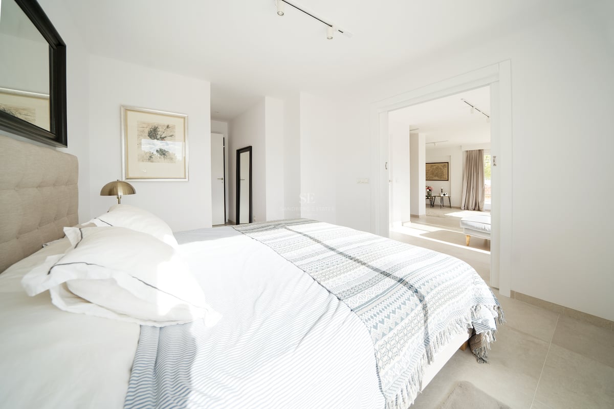 Chambre principale lumineuse avec murs blancs, carrelage grand format, tête de lit capitonnée et ouverture sur le séjour.
