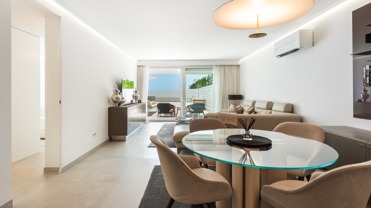 Salon lumineux avec table à manger ronde en verre, canapé crème et accès à une terrasse ensoleillée.