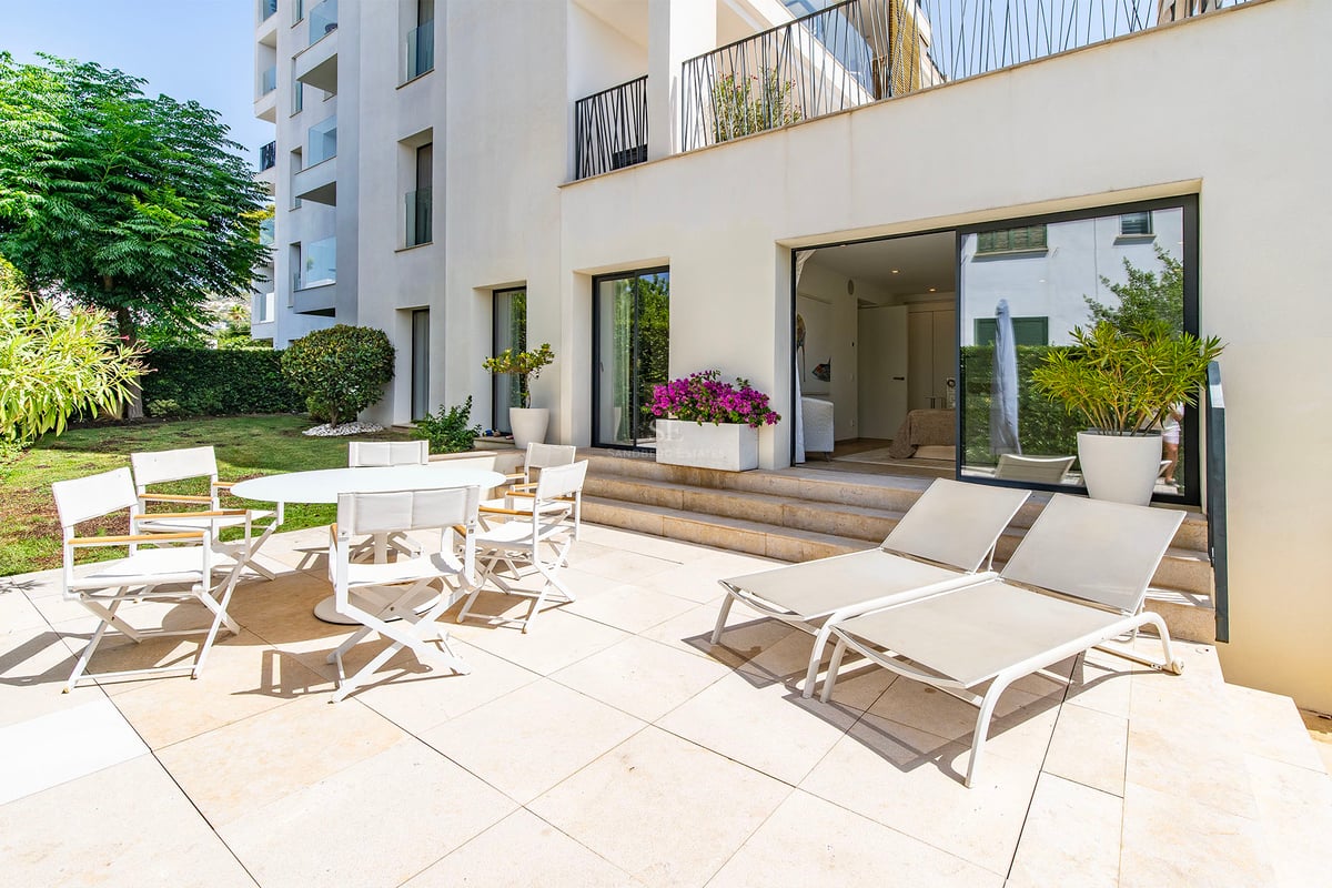 Terrasse ensoleillée avec coin repas blanc et chaises longues à côté d'un jardin et d'un immeuble moderne.