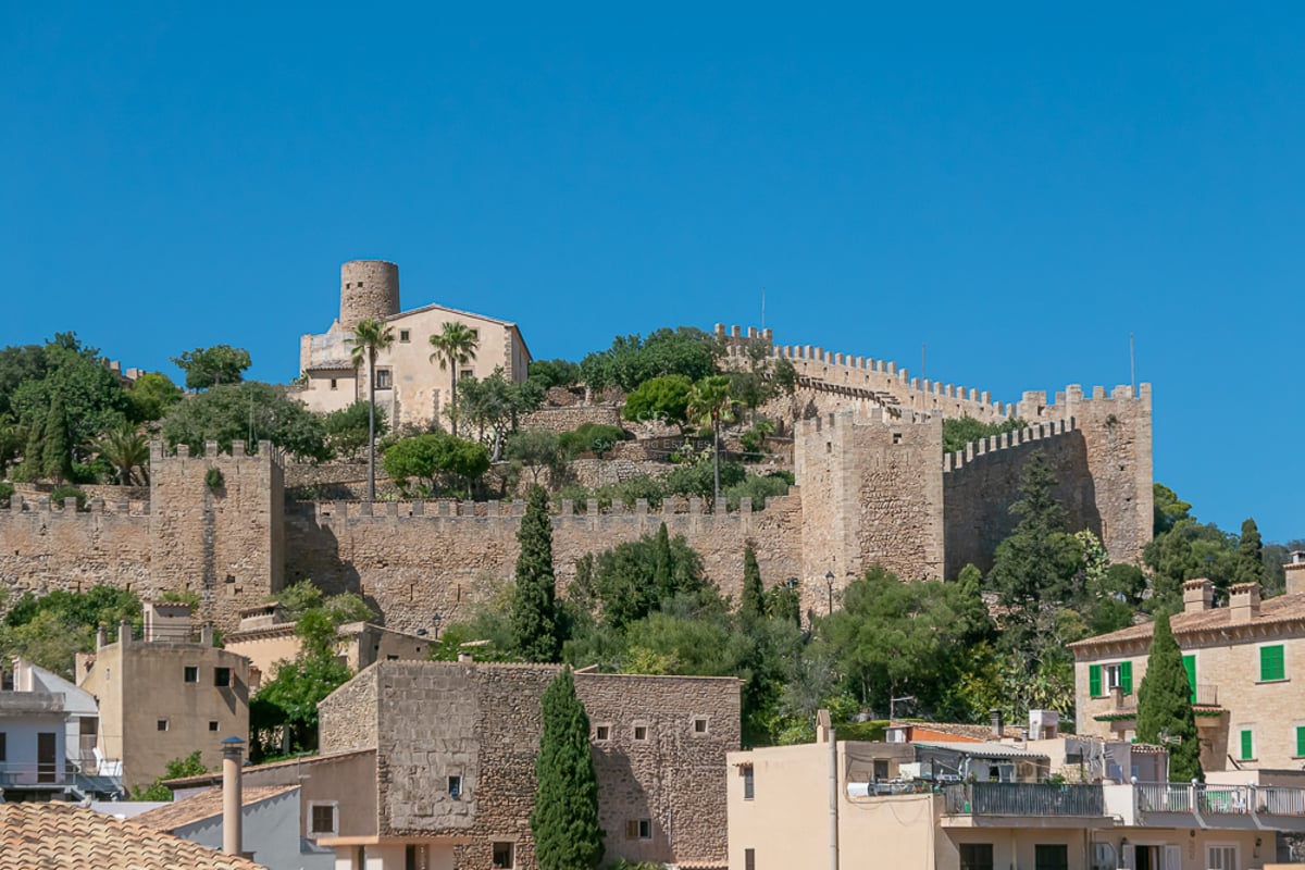 Stone walls and towers of a medieval fortress on a hill under a blue sky in a Mallorcan village.