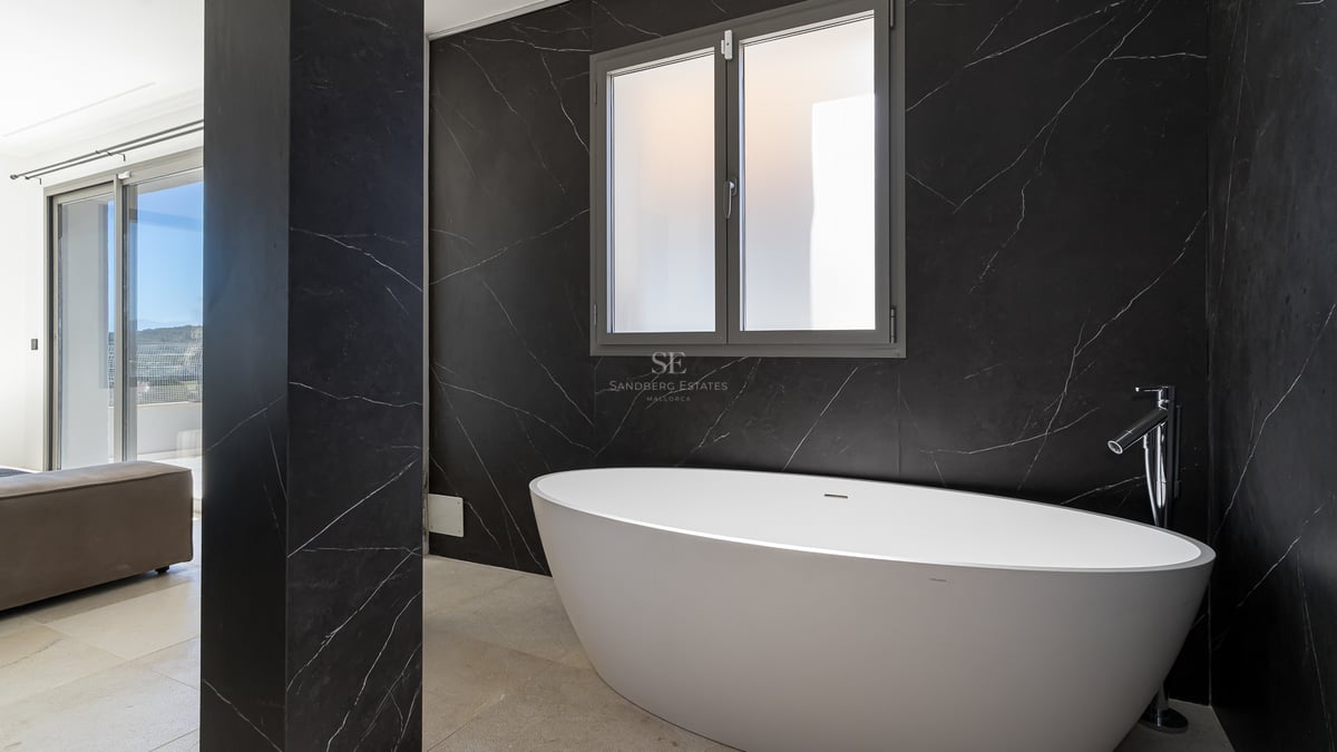 Salle de bains moderne avec baignoire ovale blanche contre un mur en marbre noir veiné et une fenêtre dépolie.