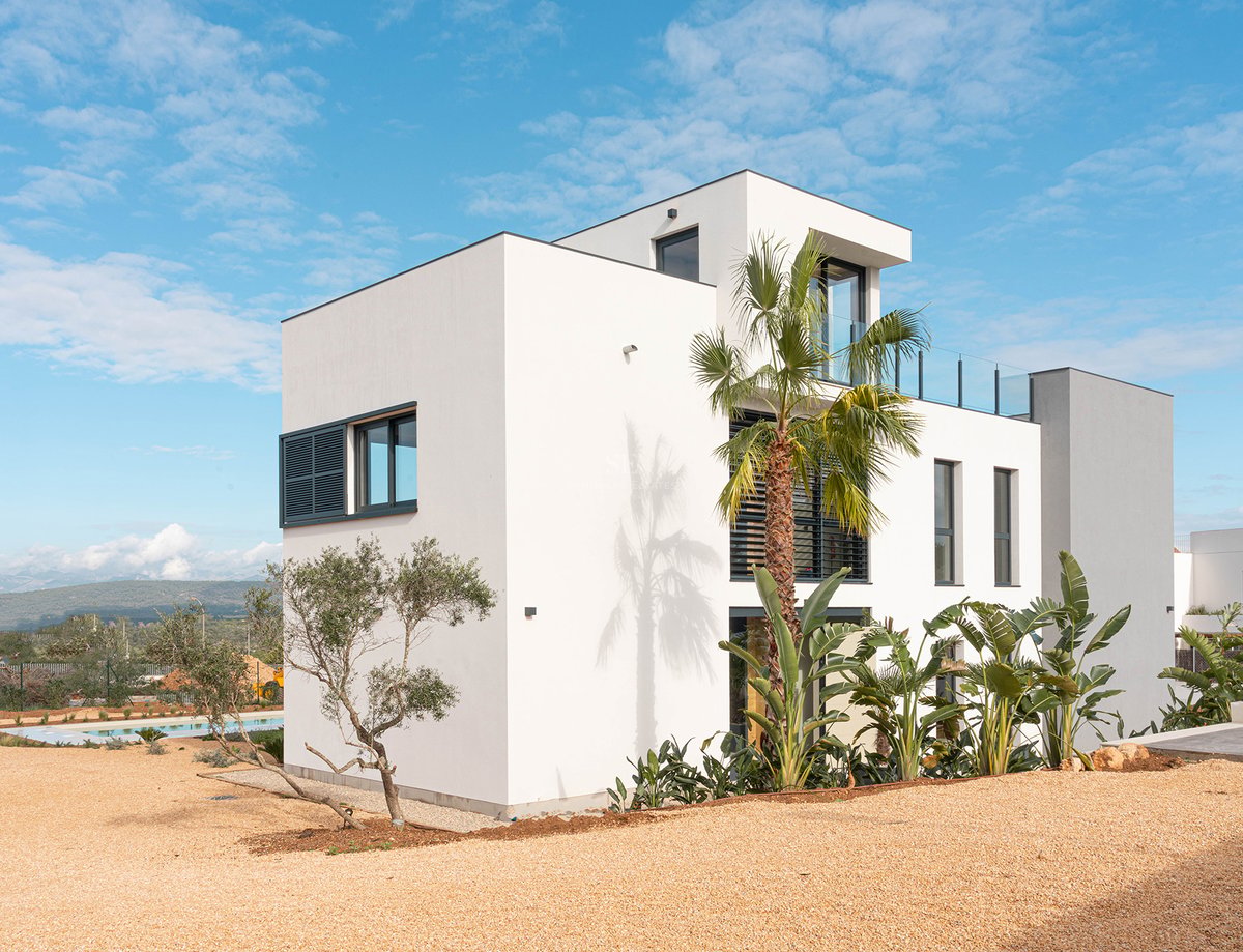 Außenansicht einer dreistöckigen weißen Villa mit moderner Architektur, Palmen und Kieshof unter blauem Himmel.