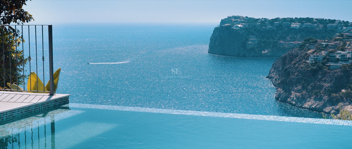 Bord d'une piscine à débordement dominant une mer bleue avec des falaises côtières et un sillage de bateau.