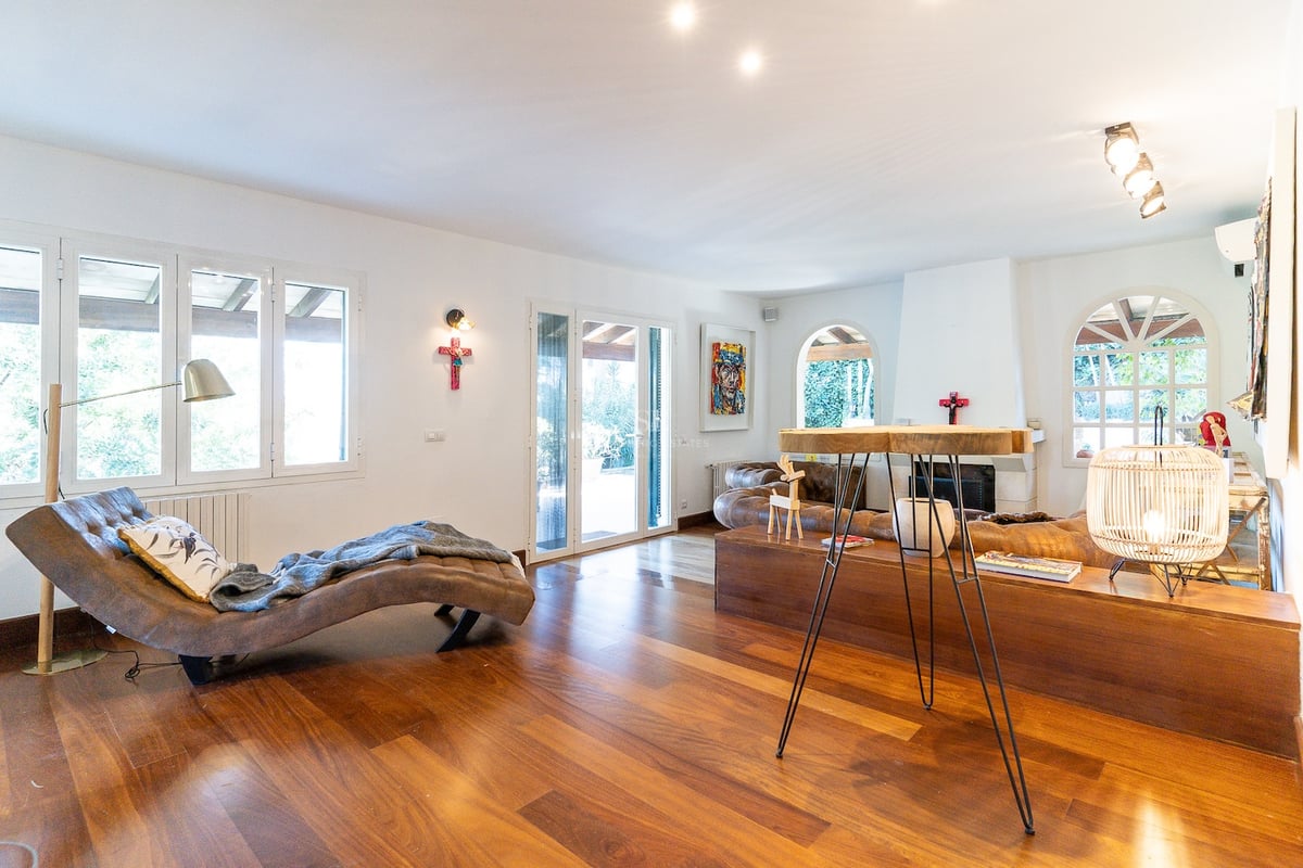 Salon lumineux avec parquet, chaise longue en cuir, canapé marron et cheminée blanche près de grandes fenêtres.