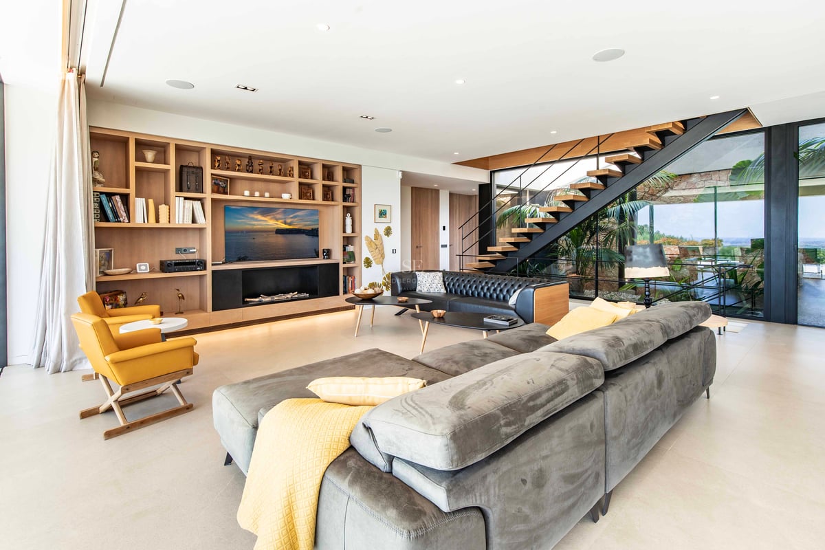 Salon moderne avec canapé d'angle gris, étagères en chêne personnalisées et escalier suspendu avec vue jardin.
