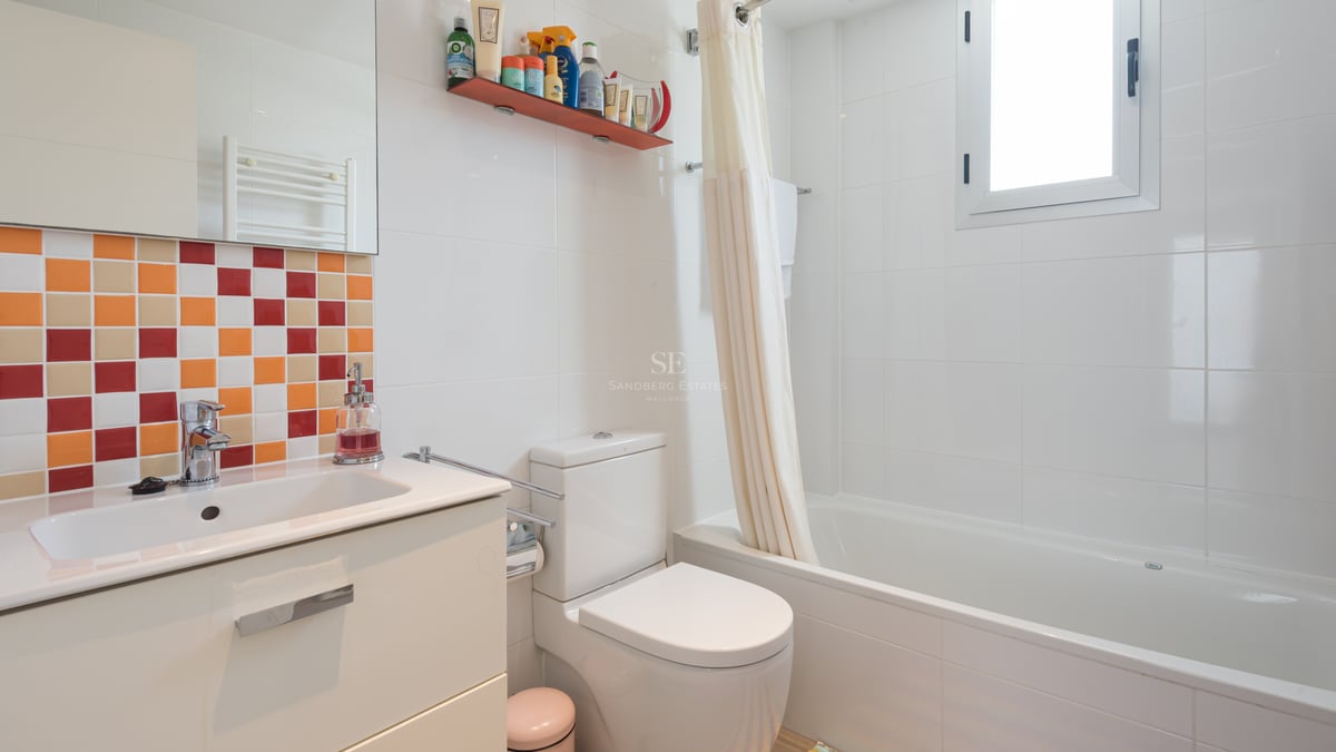 Salle de bains blanche avec mosaïques colorées, meuble vasque, toilettes et baignoire avec rideau près d'une fenêtre.