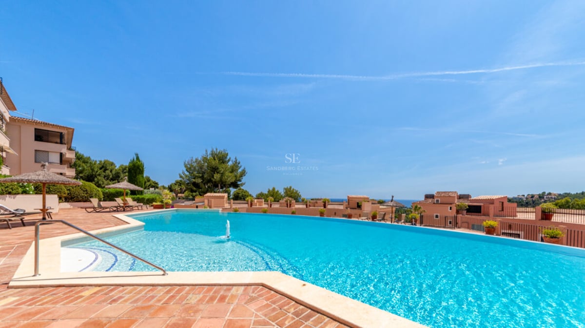 Grande piscine bleue incurvée avec fontaine, terrasse en terre cuite, parasols en paille et vue sur mer.