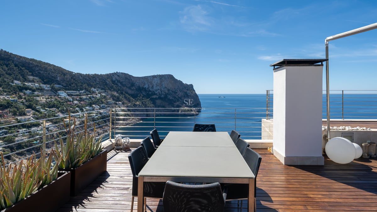 Terrasse moderne sur le toit avec une grande table à manger surplombant la mer et la côte escarpée sous un ciel bleu.