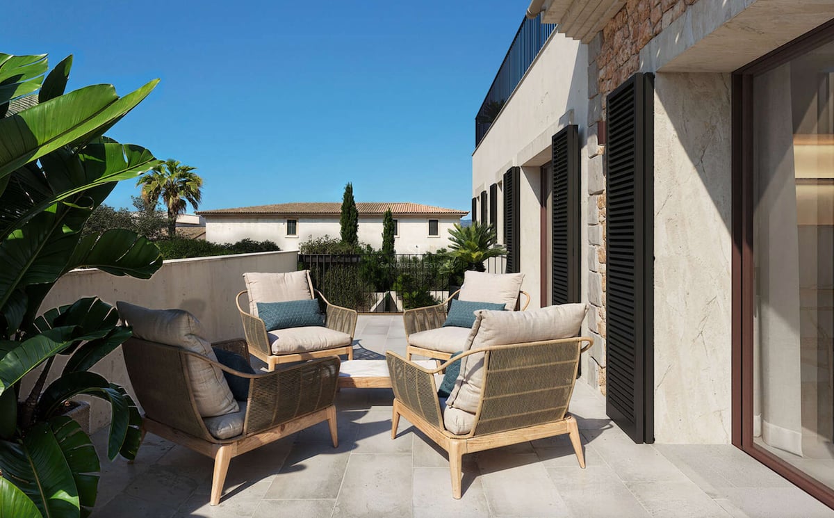 Terrasse de luxe avec quatre fauteuils tressés, coussins crème et murs en pierre naturelle avec volets.