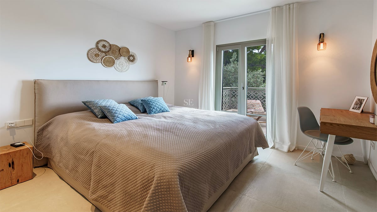 Chambre moderne avec grand lit, literie beige, coussins bleus, bureau en bois et porte vitrée donnant sur un balcon.