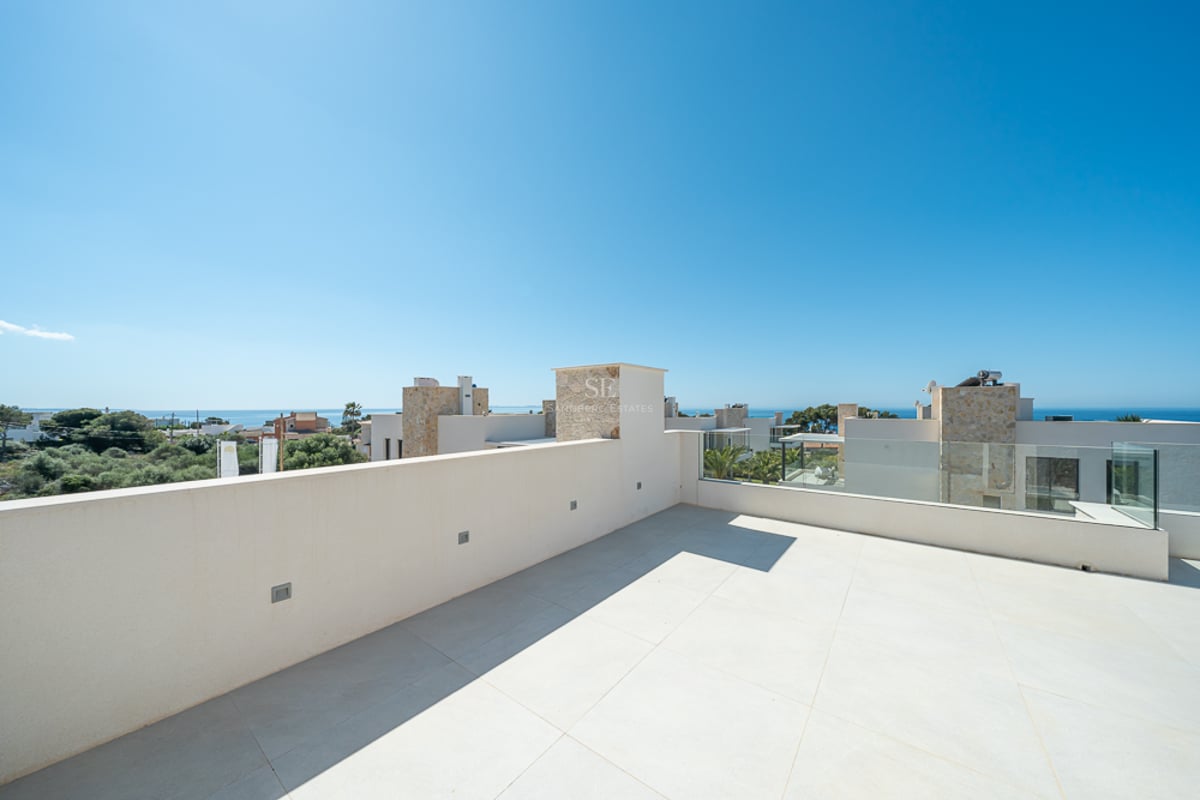 Grande terrasse de toit carrelée de blanc avec balustrade en verre, surplombant des villas modernes et l'océan.