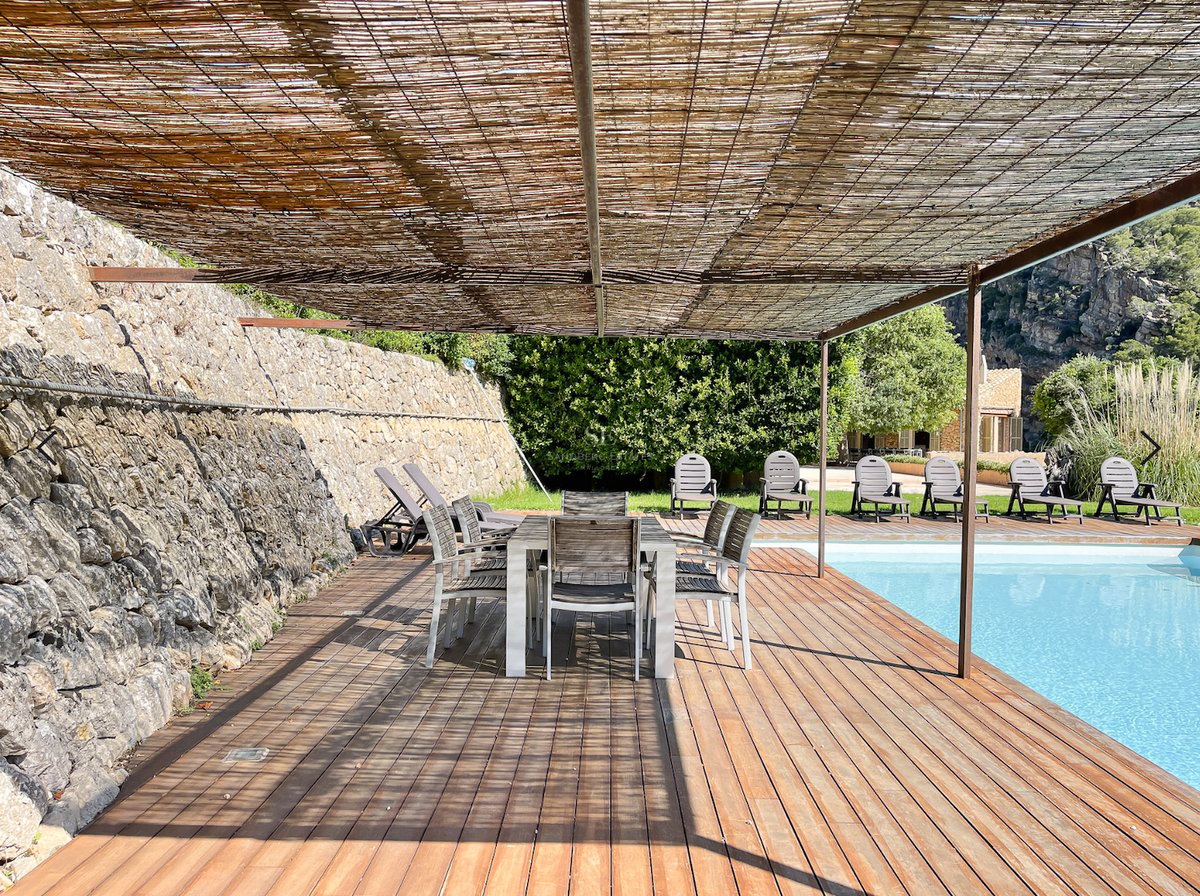 Terrasse en bois au bord d'une piscine avec table à manger sous auvent en canisse et mur en pierre.
