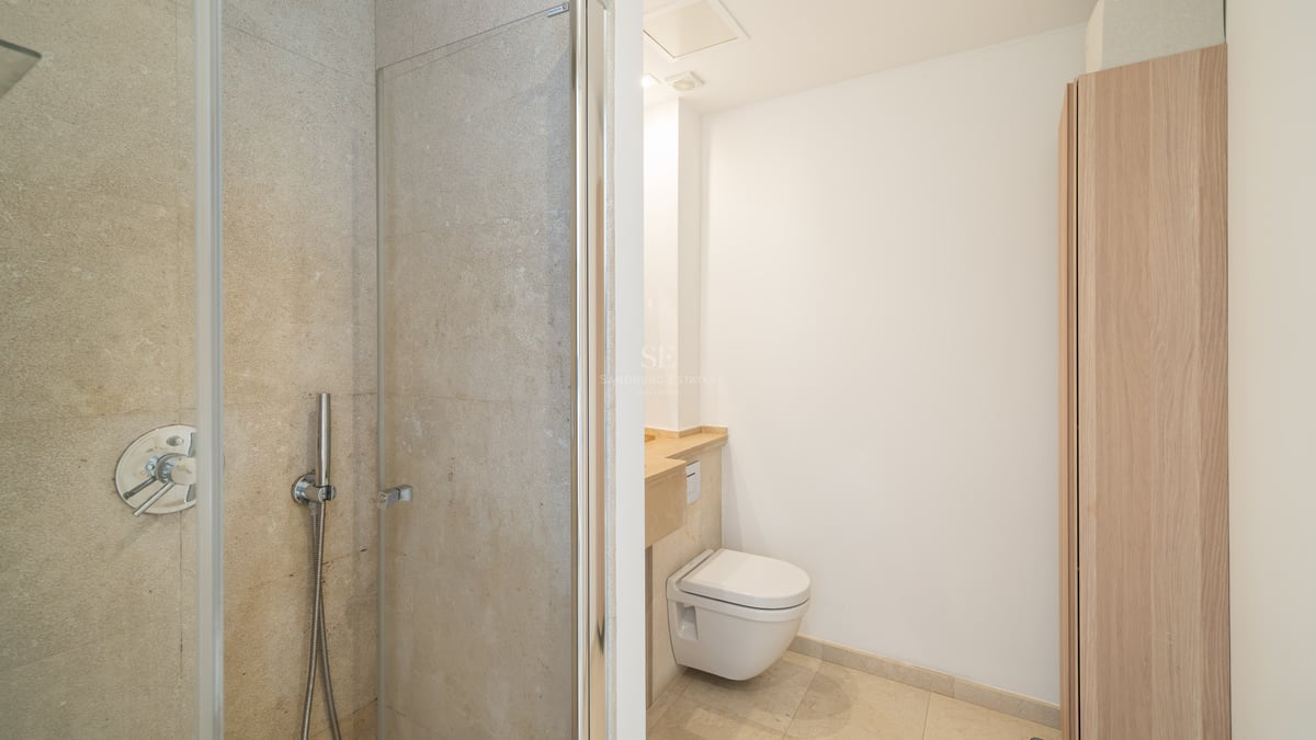 Salle de bain avec douche vitrée, toilettes suspendues et murs en pierre beige.