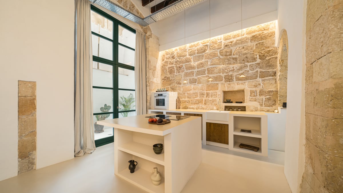 Cuisine moderne avec îlot blanc, mur en pierre rustique, hauts plafonds et grandes fenêtres.