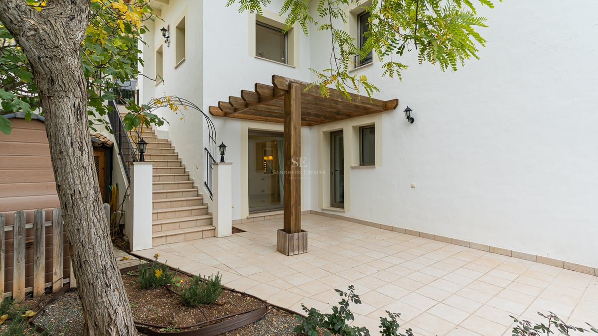 Extérieur de bâtiment blanc avec patio carrelé, pergola en bois et escaliers en pierre menant à l'étage.