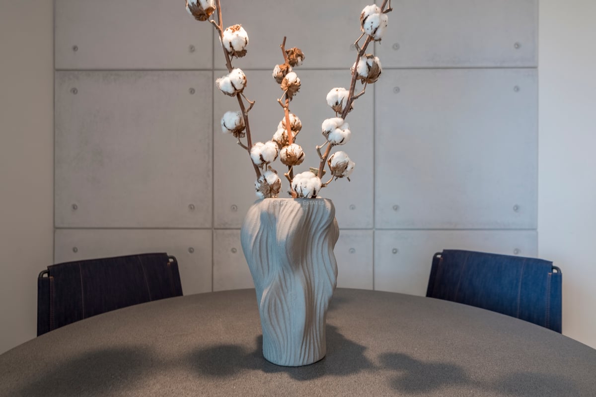 Vase gris texturé avec des branches de coton sur une table sombre devant un mur en panneaux de béton.