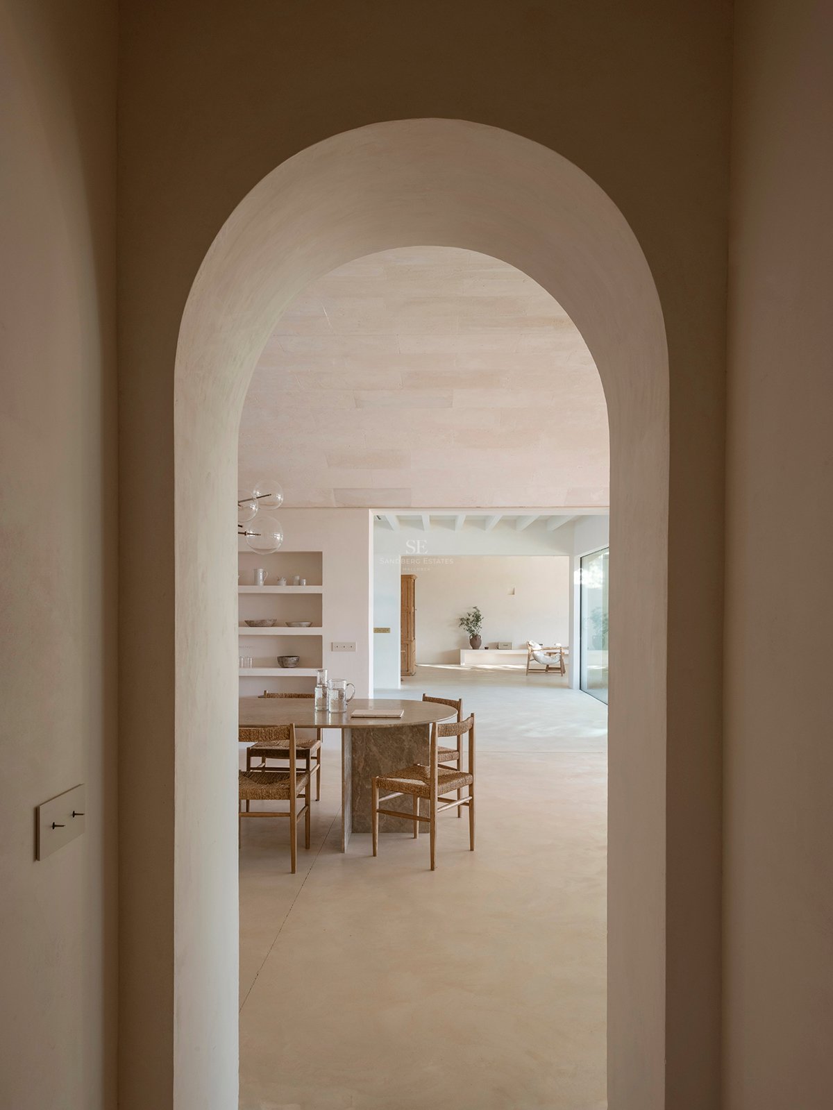 View through a plaster archway into a dining area featuring a stone table, wicker chairs, and sand-colored microcement.