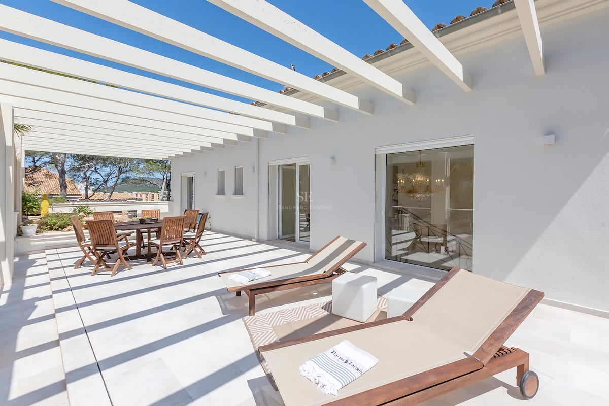 Terrasse blanche lumineuse avec pergola en bois, table à manger extérieure et chaises longues sous un ciel bleu.