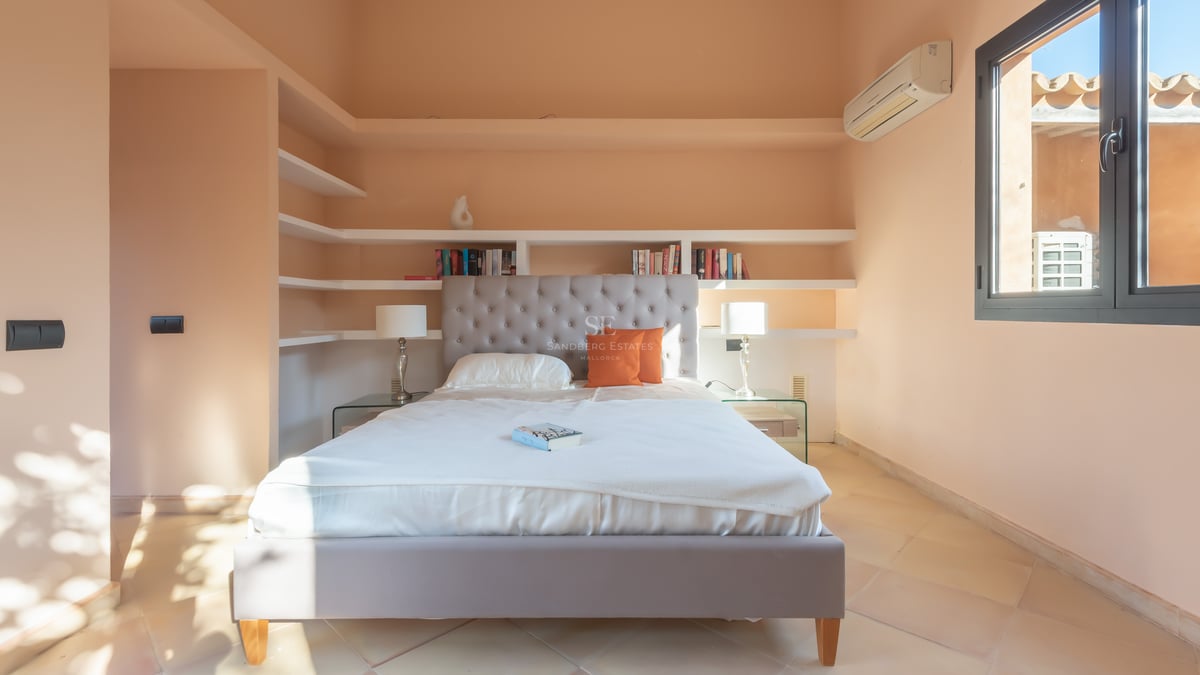 Chambre lumineuse avec tête de lit capitonnée grise, literie blanche, étagères encastrées et sol en terre cuite.