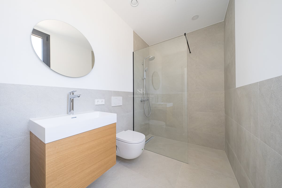 Salle de bain minimaliste avec meuble en bois clair, miroir rond, WC blanc et douche en verre avec carrelage beige.