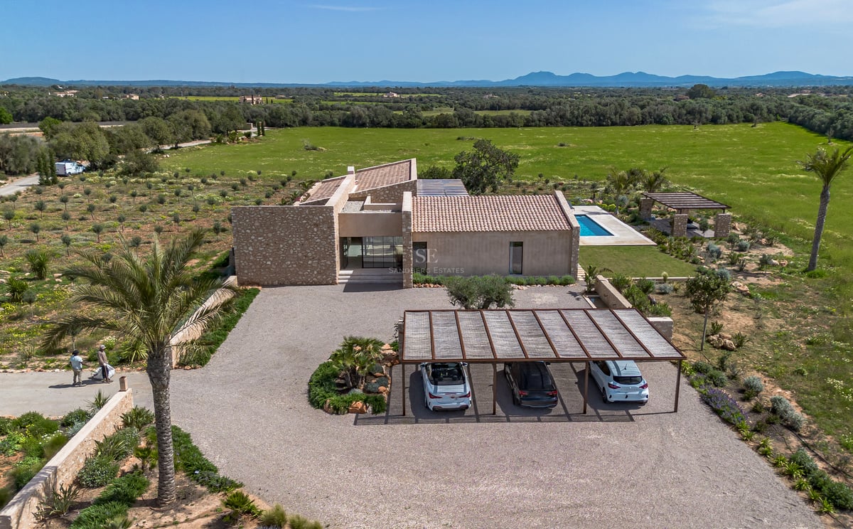 Vista aérea de una villa de piedra moderna con entrada de grava, pérgola y piscina frente al campo.