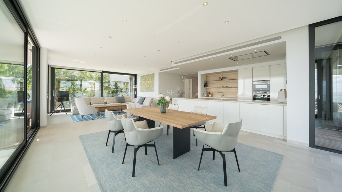Pièce lumineuse à aire ouverte avec une table à manger en bois, une cuisine blanche et des canapés près de grandes baies vitrées.